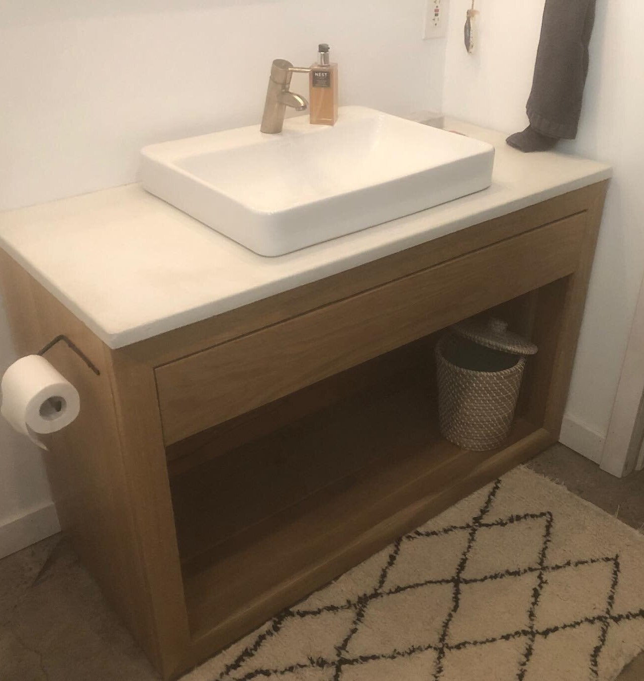 The image shows a single vanity cabinet with a concrete countertop, a sink, and a wooden drawer and shelf in a bathroom setting. The cabinet has a beige color with a white sink, and the countertop is gray.